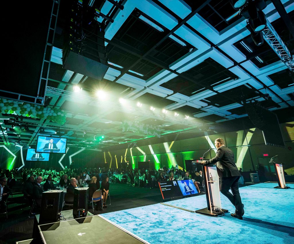 Man at lectern speaking to a crowded room at the Québec City Convention Centre. The room is set up in banquet style and screens are hanging from the ceiling.