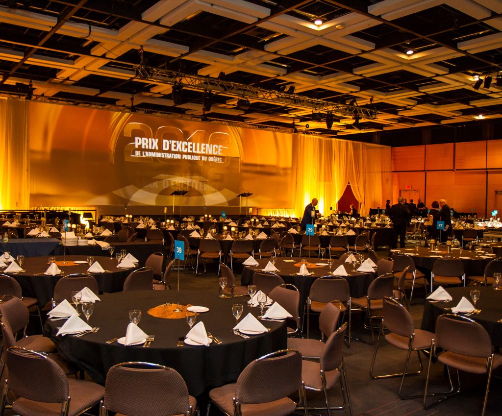 Large banquet room at the Québec City Convention Centre. Round tables with black tablecloths, yellow drapes with screen at the back of the stage.