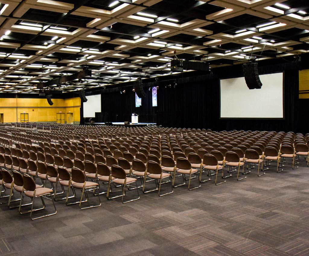 Large room at the Québec City Convention Centre with hundreds of chairs, theater style. Two screens, a small stage and lectern are located at the front of the room.