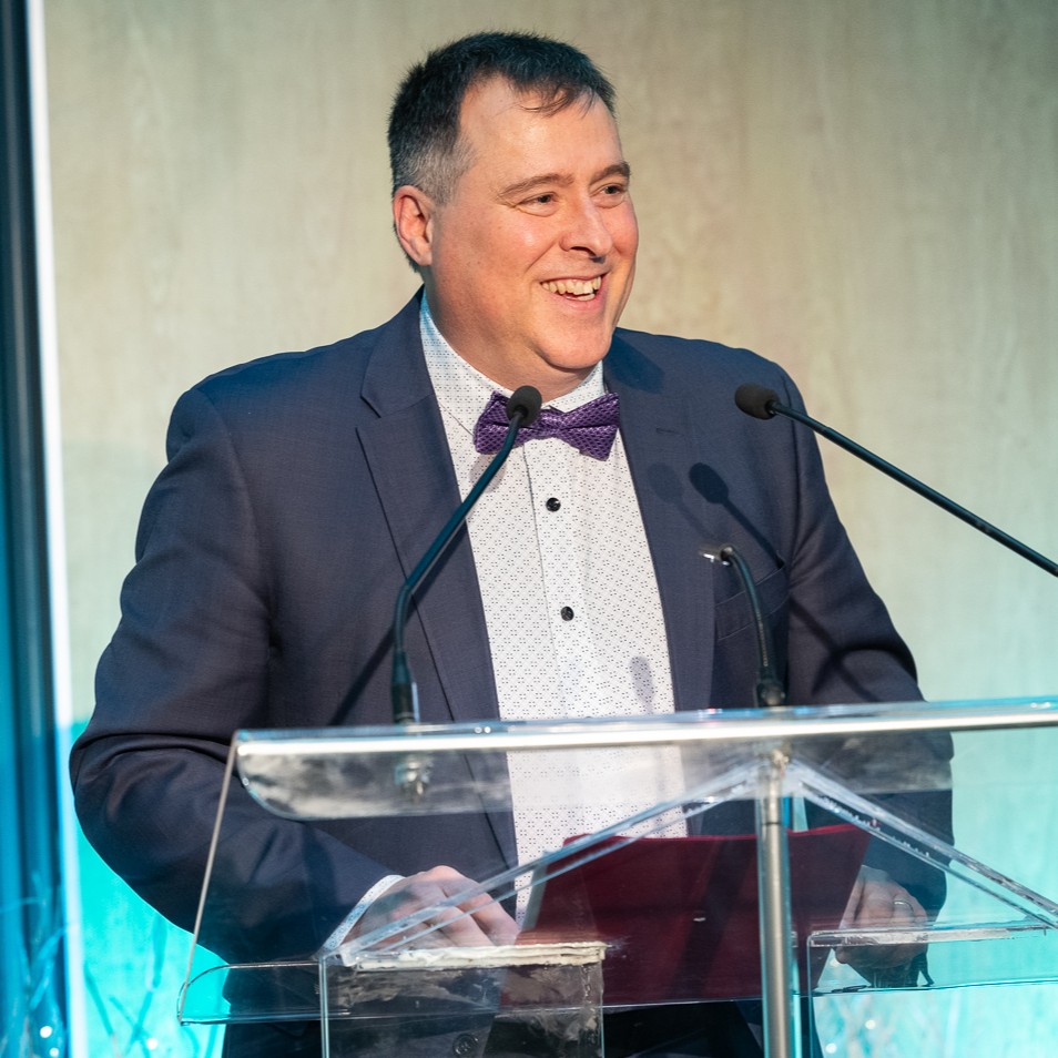 Ambassador and researcher Luc Beaulieu speaking at the microphone in front of a podium during the presentation of his “Ambassador of the Year 2024” award at the Hommage aux ambassadeurs evening.