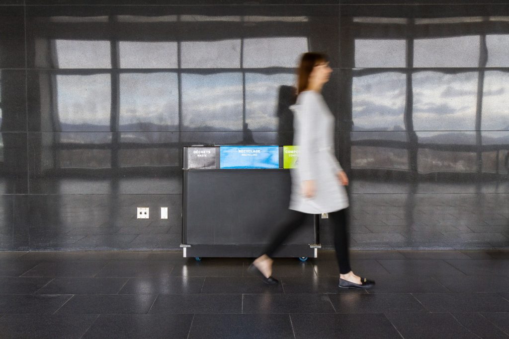 Femme passant devant un bac, nommé ilôt multimatière. Il permet d'avoir accès à une poubelle, un bac de recyclage et de compost au même endroit.