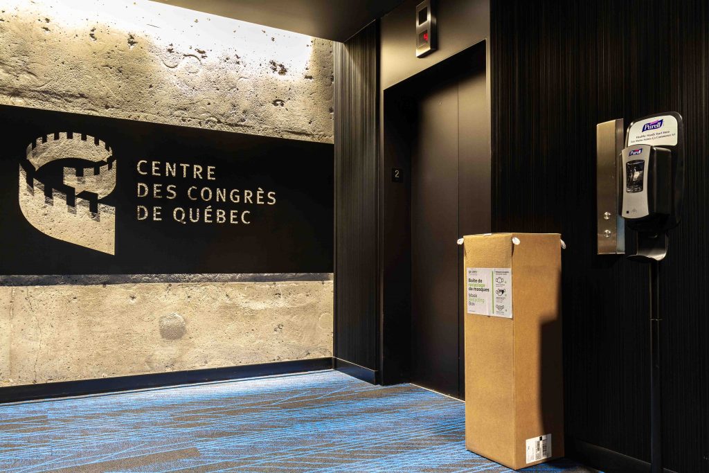 Entrée de l'étage administratif du Centre des congrès. On y voit un ascenseur, le logo du Centre, une distributrice de désinfectant à main ainsi qu'une boite de récupération de masques jetables lors de la Covid-19.