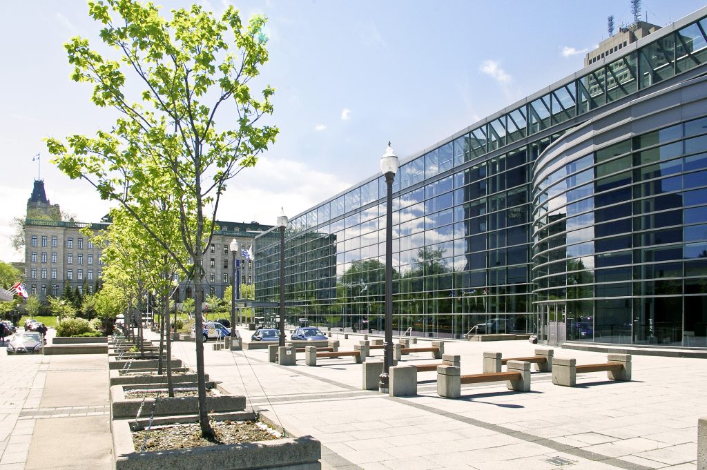 Façade extérieure du Centre des congrès et de la Promenade Desjardins au printemps, avec un arbre verdoyant et vue sur le Parlement