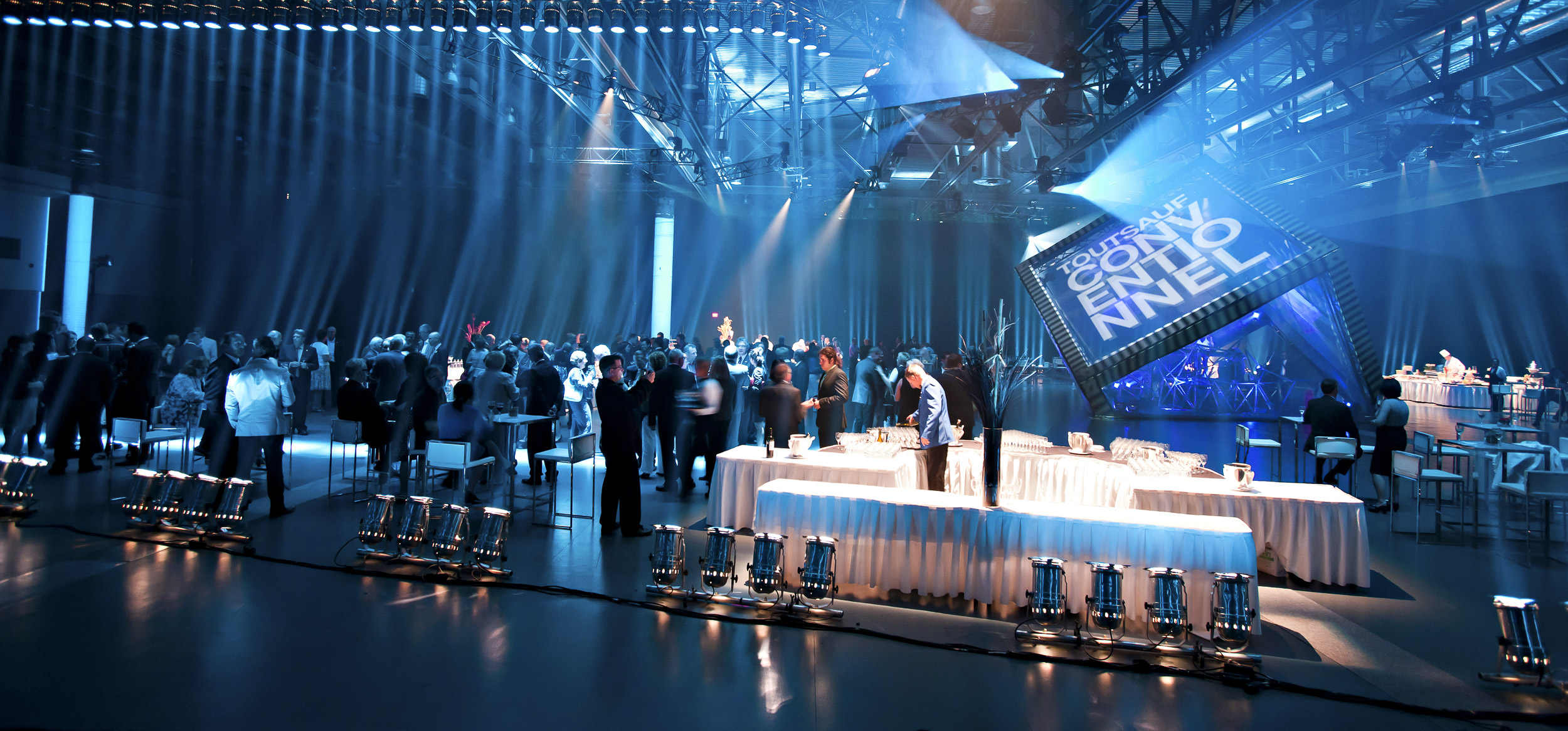 Salle 400 toute éclairée en bleu avec un grand cube au milieu de la salle. On y voit les tables du traiteur Capital HRS.