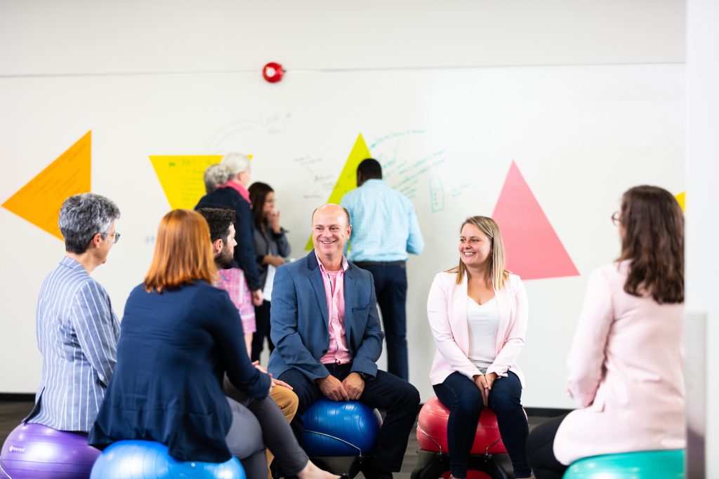 A team of delegates in room 309, sitting on balance ball chairs with smiles on their faces. Behind them, another team is writing their ideas on walls decorated with colorful triangle shapes.