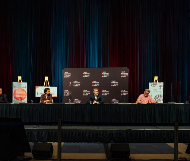 Table set up on a stage, with 3 men sitting with microphones during a press conference for the Salon du livre at the Québec City Convention Centre. 