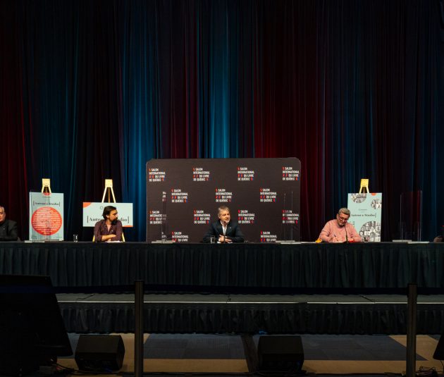 Table set up on a stage, with 3 men sitting with microphones during a press conference for the Salon du livre at the Québec City Convention Centre.