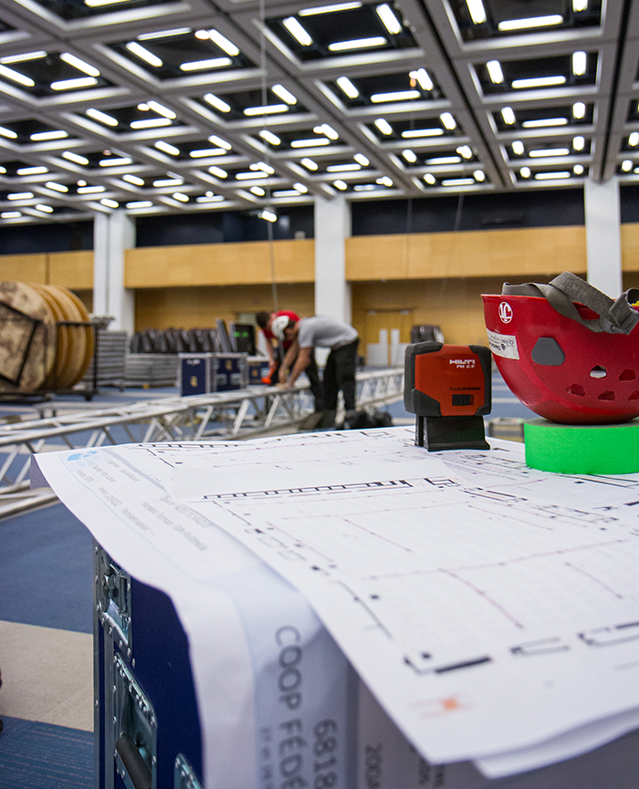 Salle en montage au Centre des congrès de Québec où l'on peut voir des gréeurs en train d’installer de l’équipement. Un casque de sécurité et des gants sont posés sur une caisse de matériel.