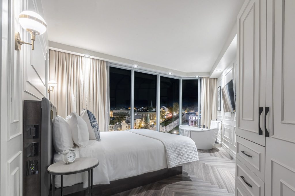 Chambre de l'Hôtel Le Capitole à Québec avec grand lit et literie blanche. Bain dans le coin de la chambre avec vue sur Place d'Youville.