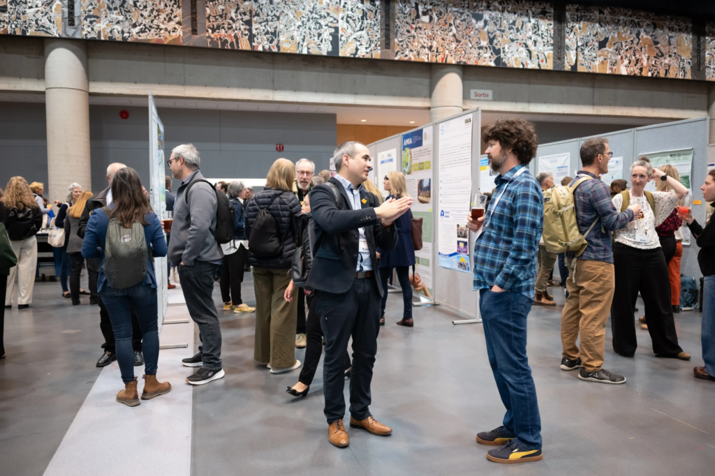 Activité de type "Poster Session" se déroulant dans la salle 400 lors du congrès international IAIA26. On peut voir des gens debout qui discutent et, en arrière-plan, l'immense murale Paraiso, l'une des 4 oeuvres d'art emblématique du Centre des congrès de Québec.