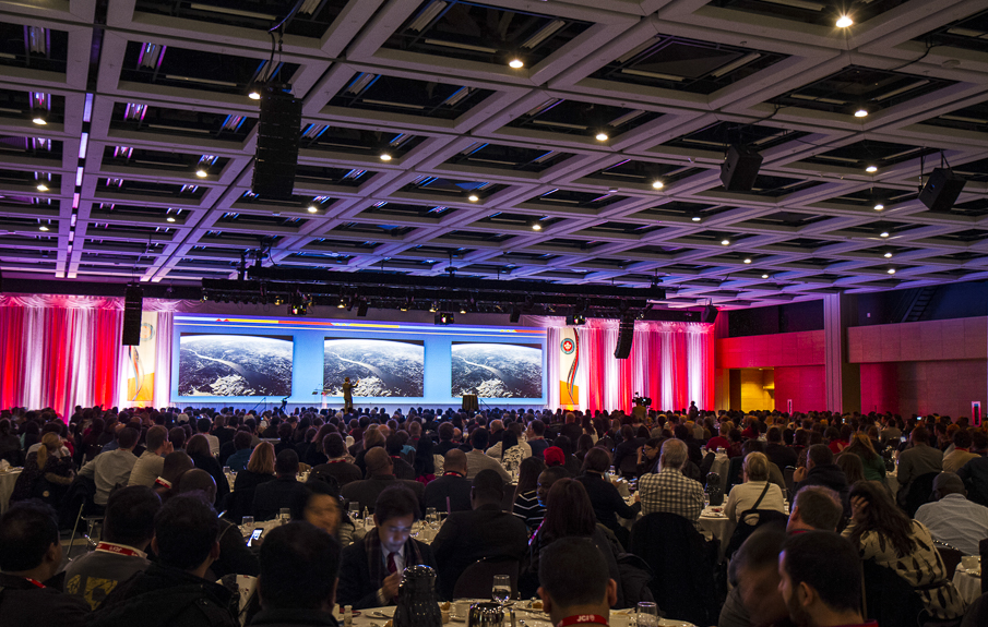 Grande salle remplie de gens assis à leur table ronde de banquet avec une scène en arrière plan, éclairée en bleu, lors de l'événement JCI 2016 à Québec.