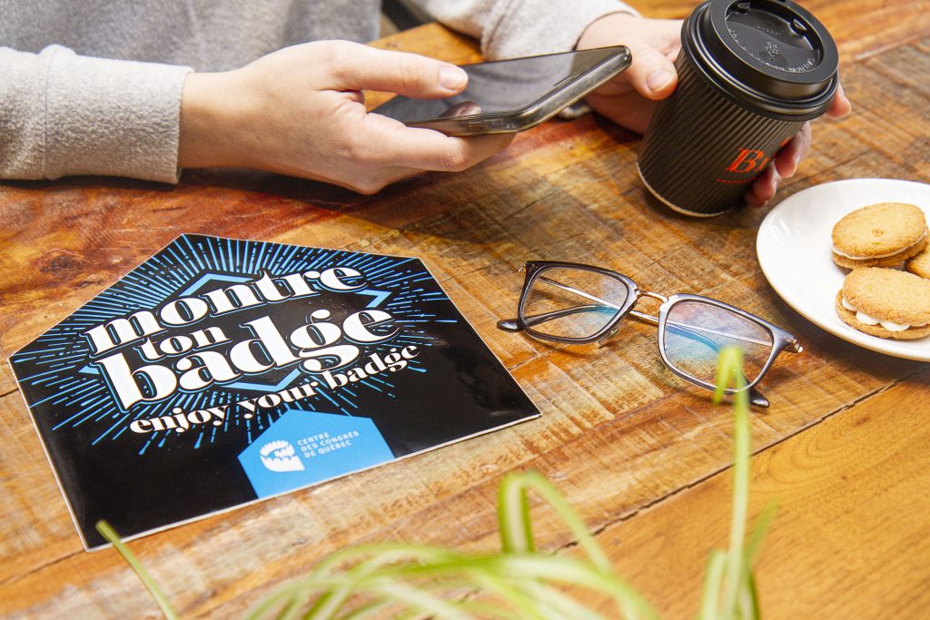 Close up on table with a woman's hand holding a phone and a coffee cup. Enjoy your badge sticker with glasses and cookies are resting on the table.