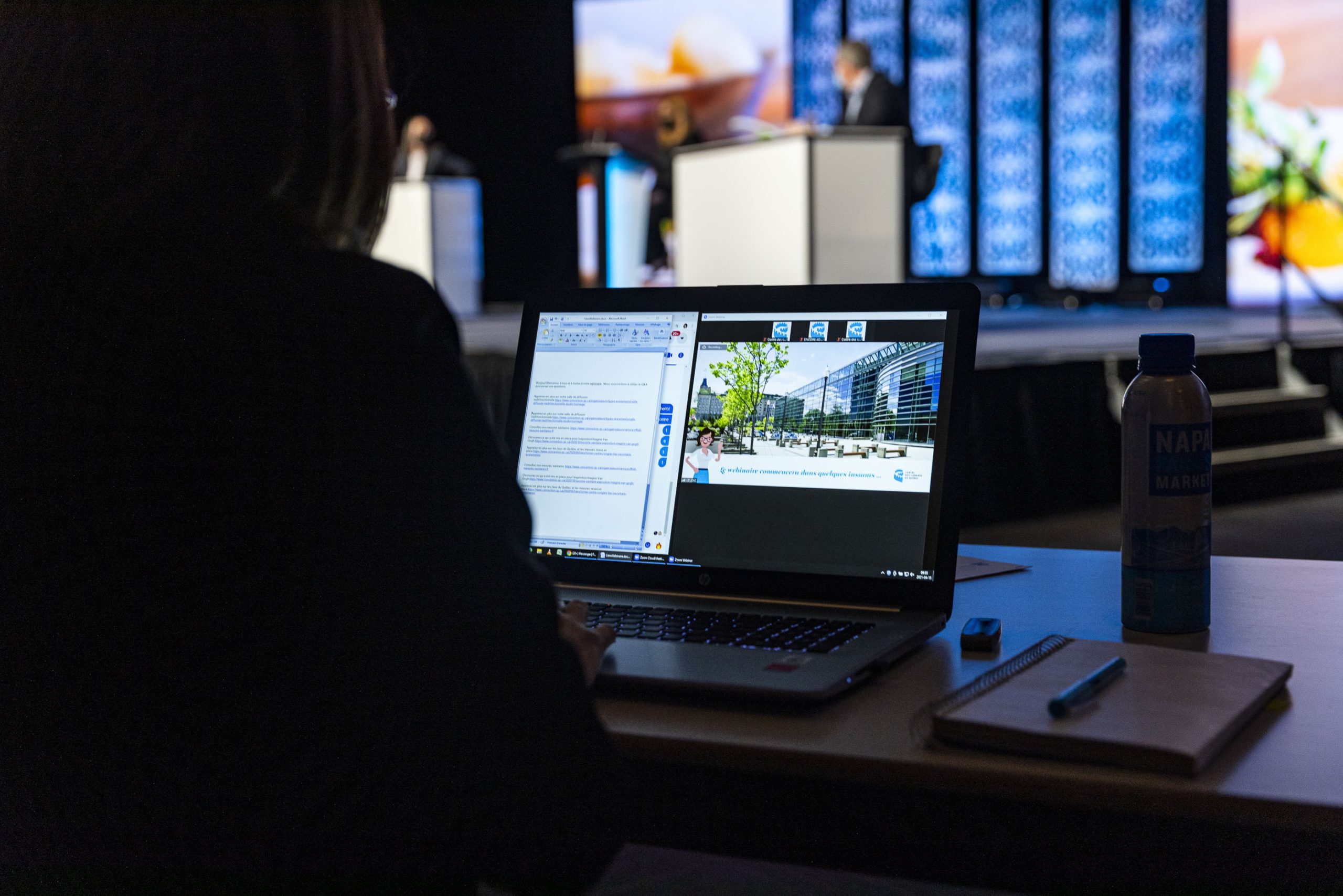 Women seated at a desk with computer on table, watching a stage with 3 people presenting.