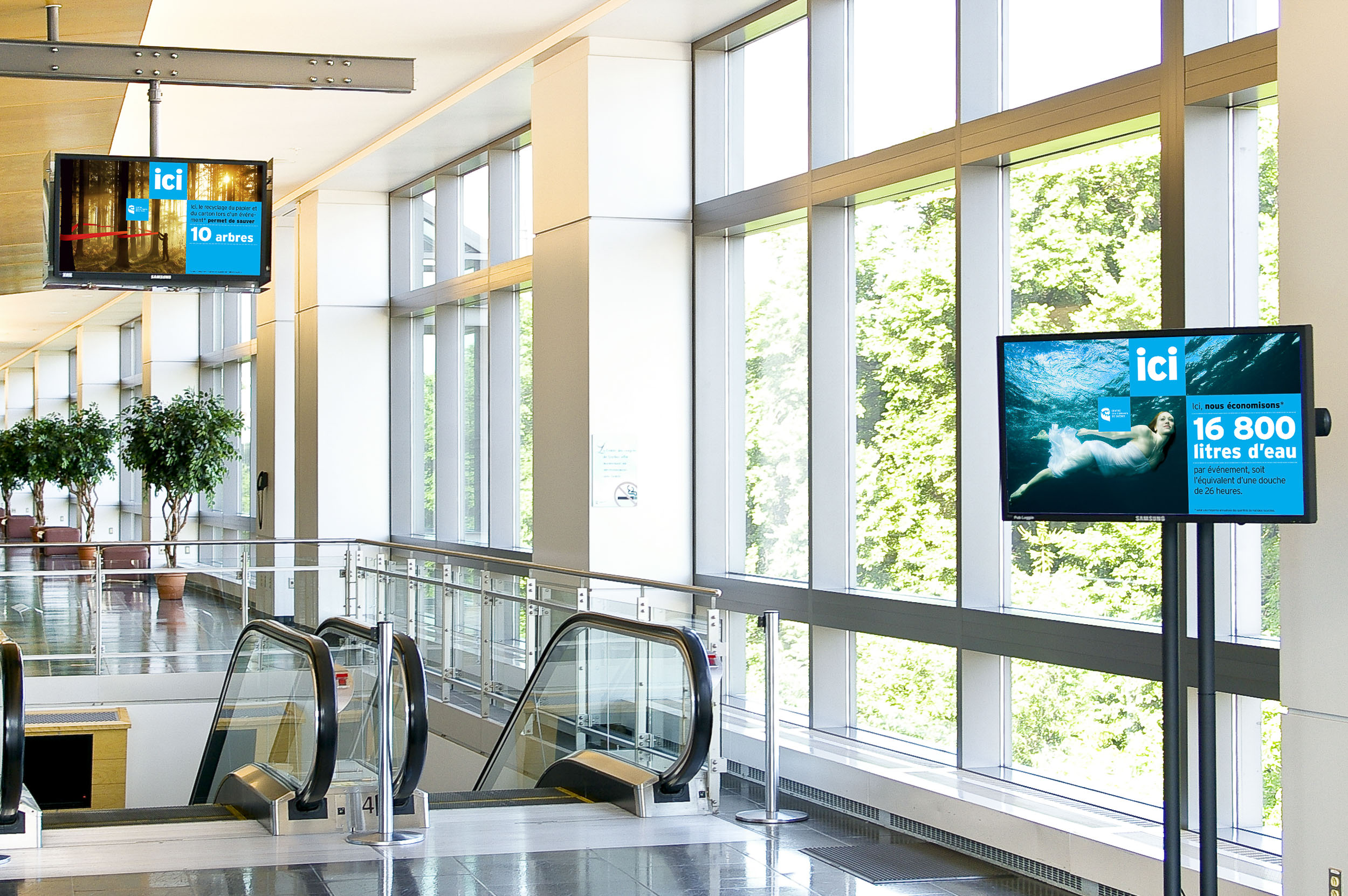 Hallway with large windows and escalator, next to 2 digital signage screens.