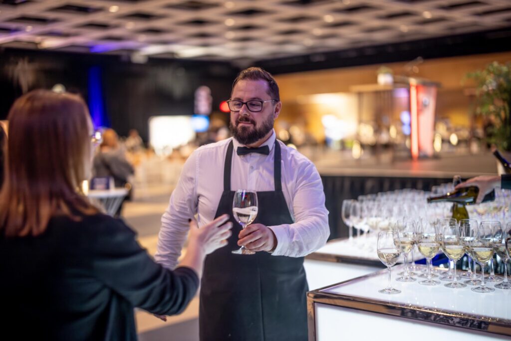 Serveur de Maestro Culinaire offrant un verre de vin à une dame dans la salle 200 lors de l'événement Soirée Exquise 2024.