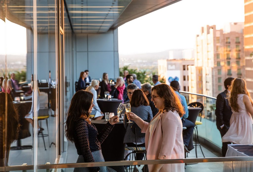 2 jeunes femmes avc un verre de vin à la main, avec d'autres congressistes sur la terrasse de l'espace urbain. Ils sont assis autour des petites tables cocktail.