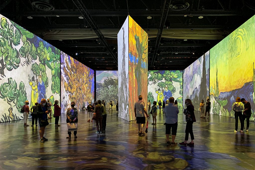 Salle 400 transformée par l'exposition immersive Imagine Van Gogh. Des gens circulent à travers les structures colorées par les projections de Van Gogh.