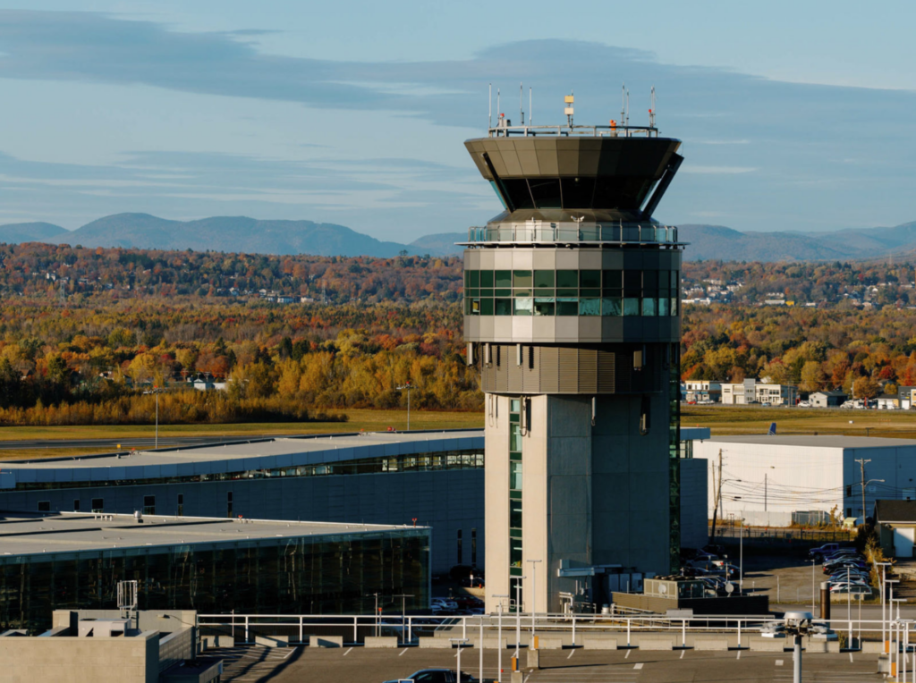 Vue aérienne de la tour de contrôle de l'aéroport international Jean-Lesage de Québec avec des montagnes aux couleurs de l'automne en arrière-plan et un ciel bleu