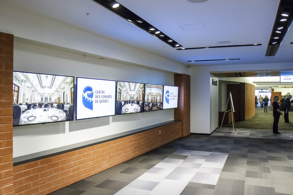 Écrans aux murs du niveau 2 du Centre des congrès de Québec montrant le logo et les salles. 