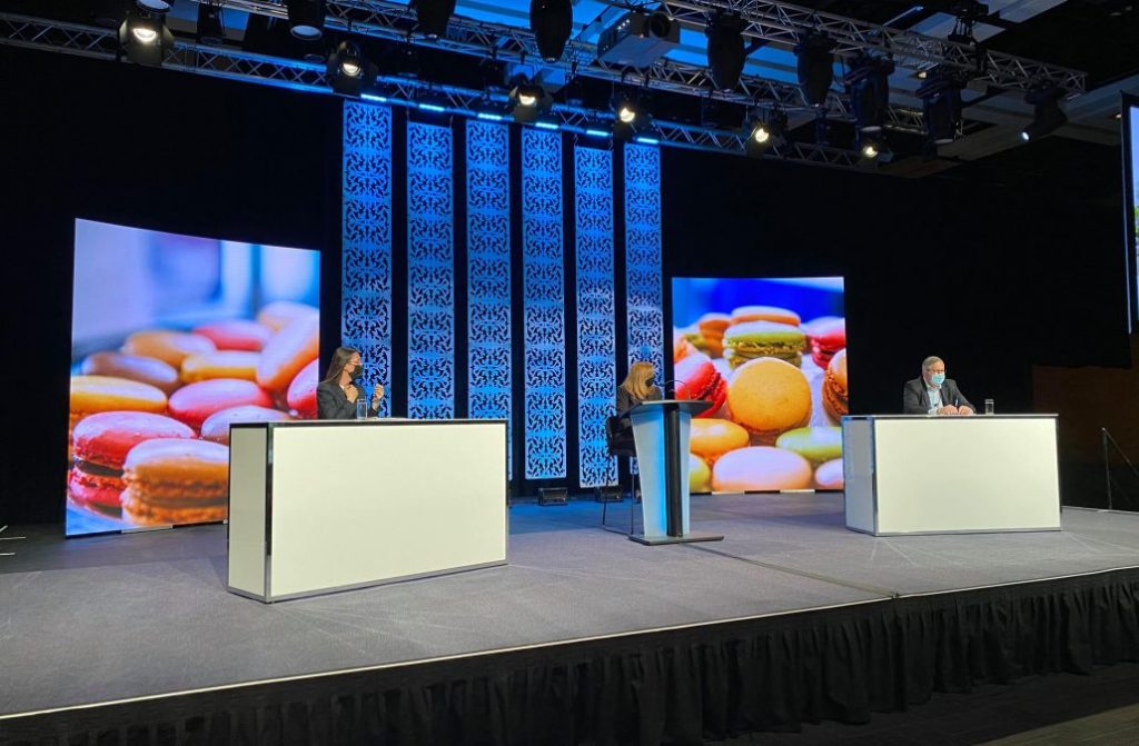 A women is seated at a lectern and is talking to a men and a women during a virtual event at the Convention Centre.