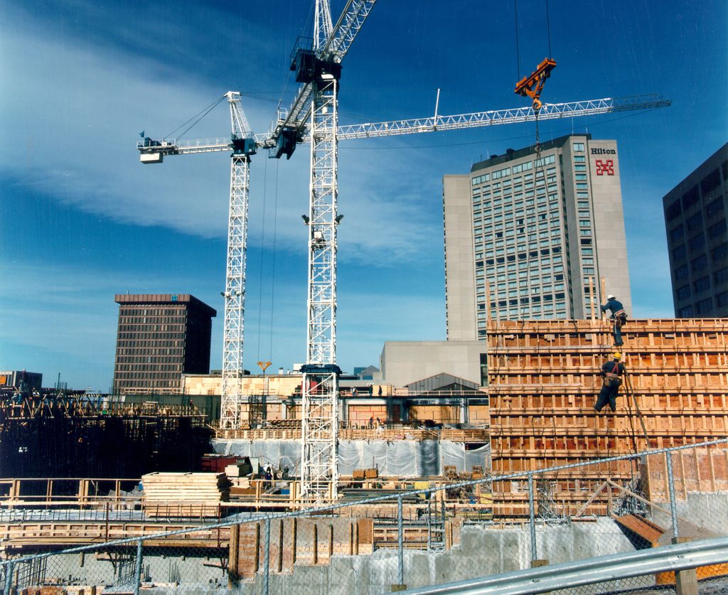 Chantier de construction du Centre des congrès de Québec, avec 2 grues, le Hilton au loin, et une structure de bois où 2 travailleurs grimpent.