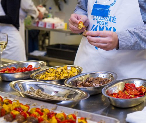 Multiple aluminium bowls with colorful food inside. A person wearing an apron is standing nearby.