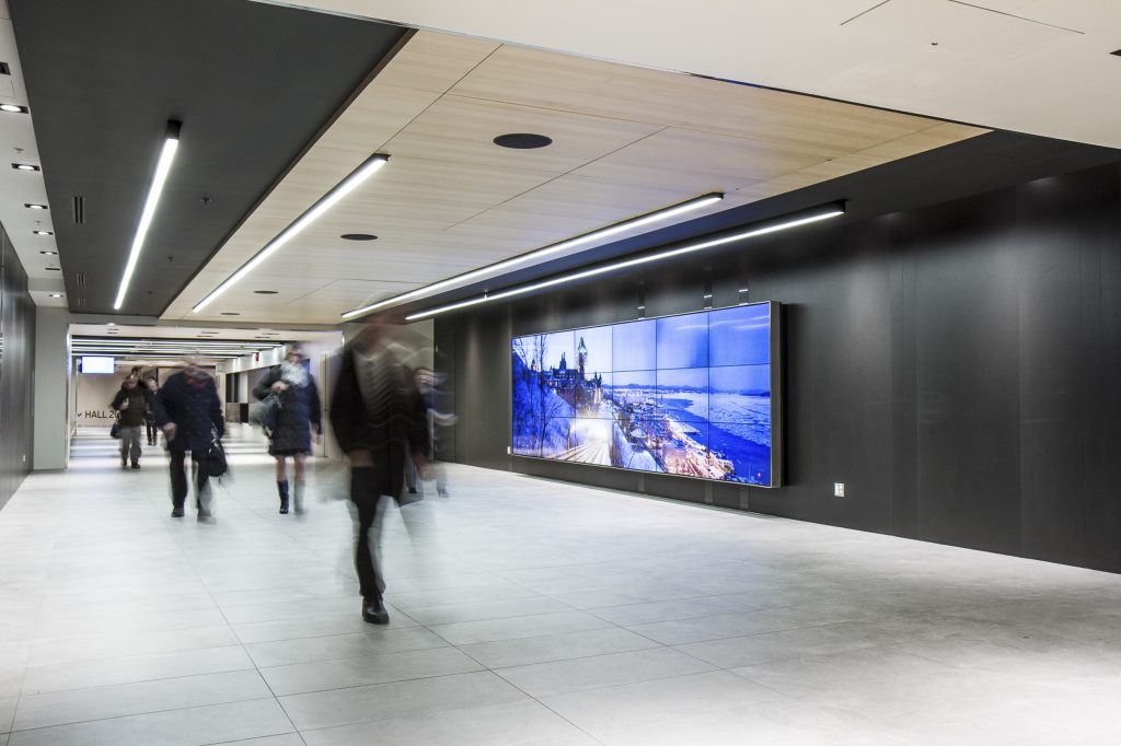 Public hallway at the Québec City Convention Centre with people walking about. Large video walls is on and showcasing the city.