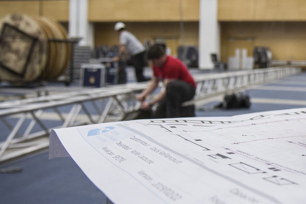 Un plan de salle en premier plan, et deux hommes qui travaillent au montage de la salle sur des ponts d'éclairage et du matériel audiovisuel.